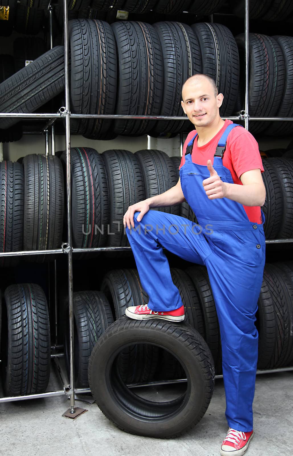 A smiling mechanic in a garage standing next to a rack full of tires and making a positive gesture.