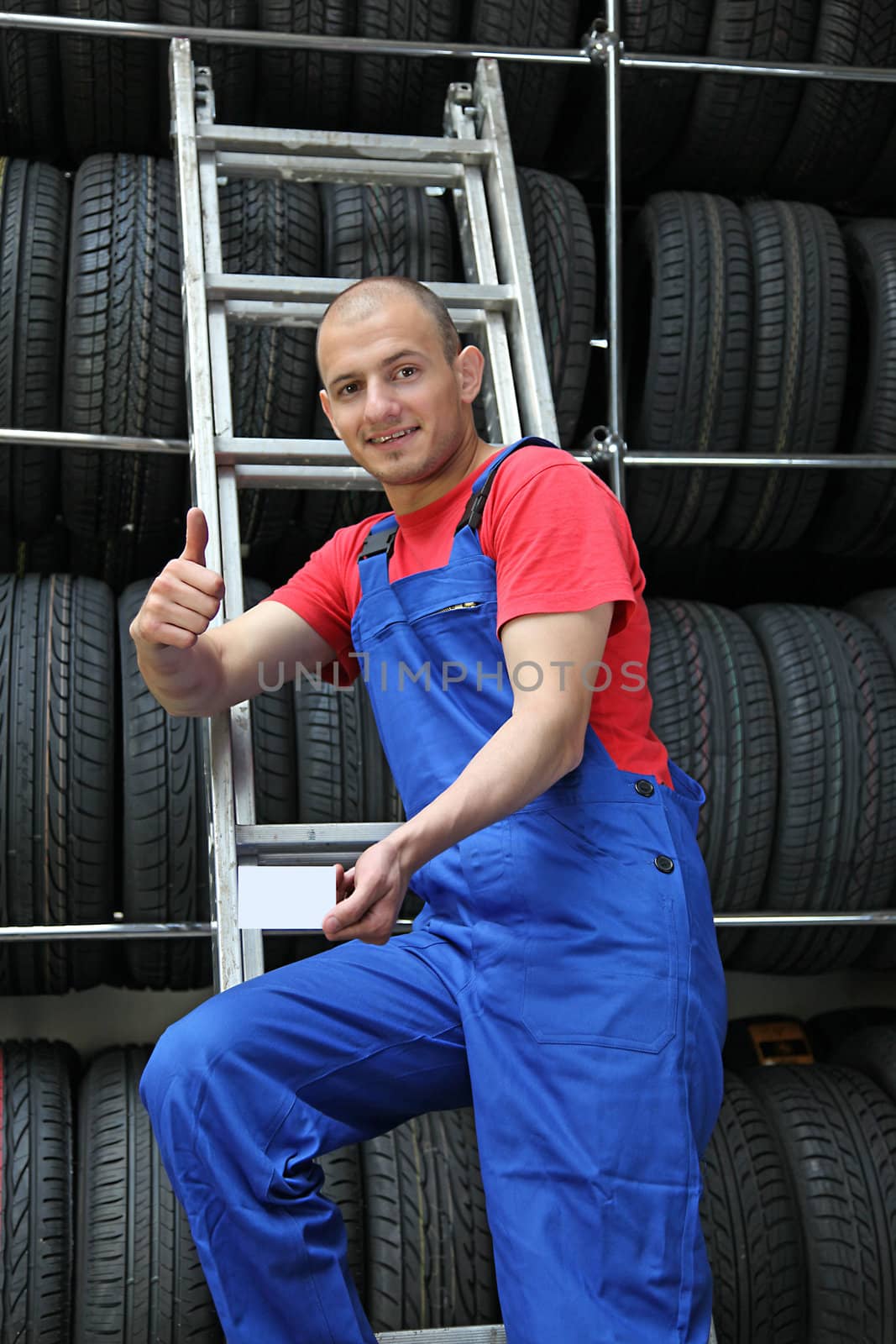 A smiling mechanic in a garage standing on a ladder next to a rack full of tires and making a positive gesture.