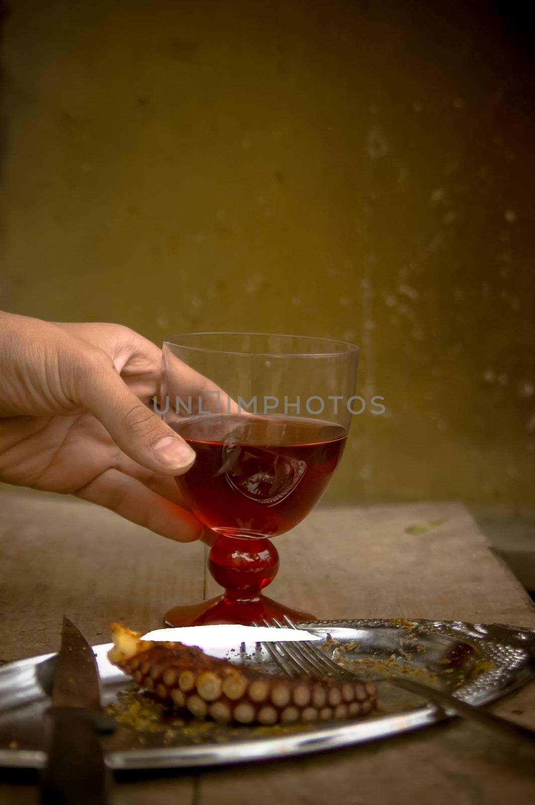 A hand holding a glass with red wine and a plate with barbecued octopus piece