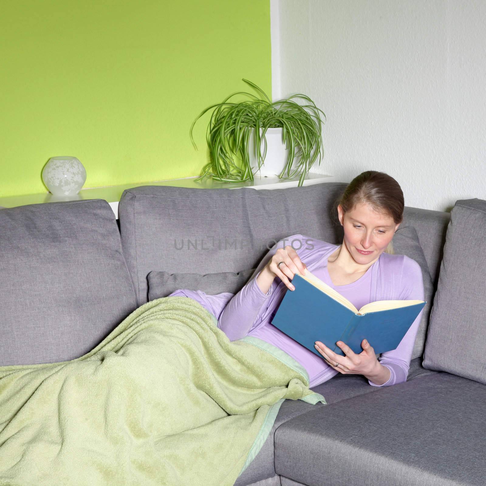 Attractive mature woman lying relaxing on a sofa in her living room with a blanket over her legs reading a book
