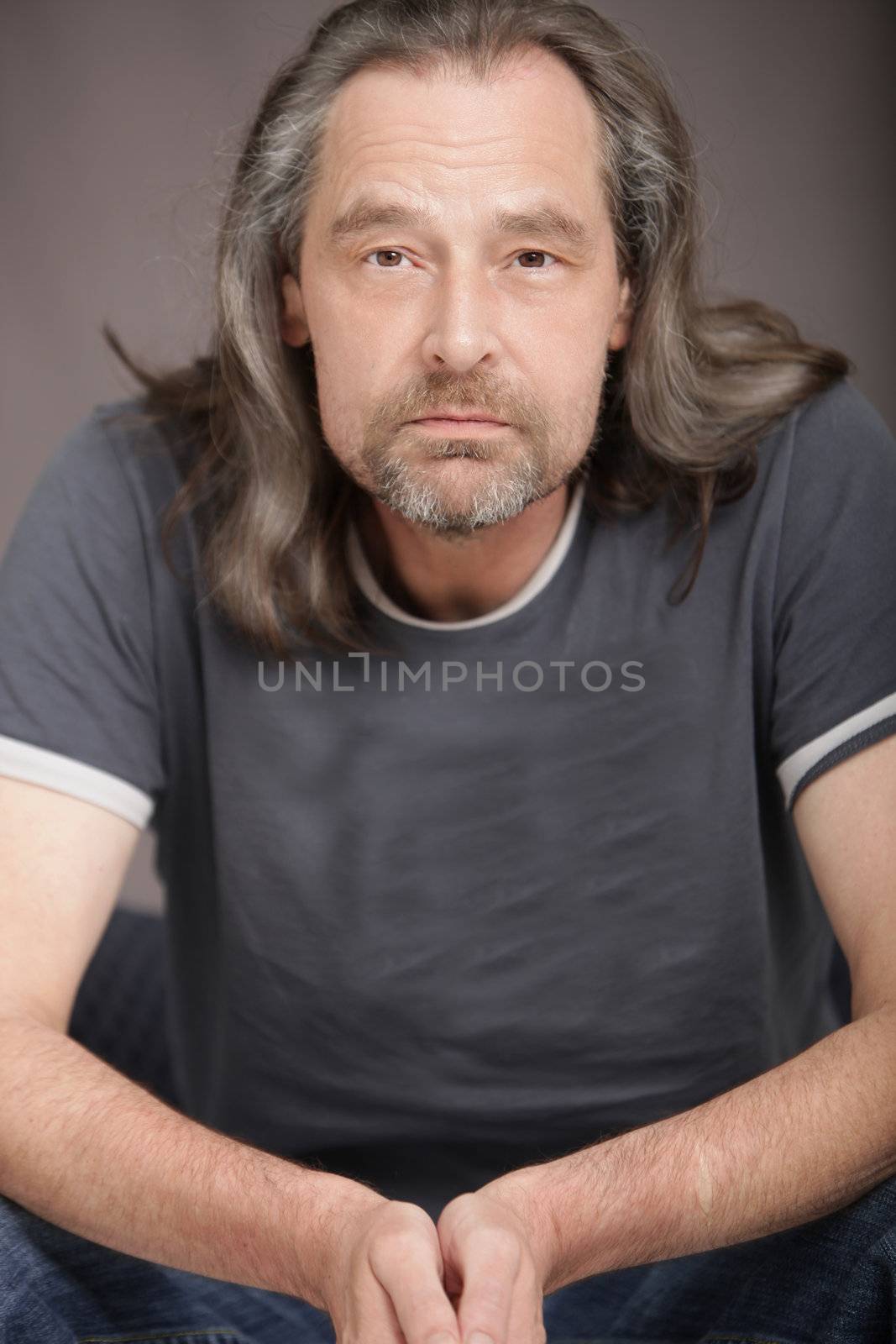 Attractive middle-aged man with a beard and long hair sitting in a chair facing the camera with a serious expression
