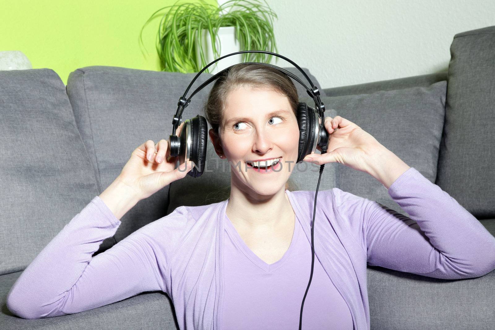 Laughing mature woman listening to music sitting on a couch in her living room wearing a pair of headphones and looking sideways to the left of the frame while facing the camera