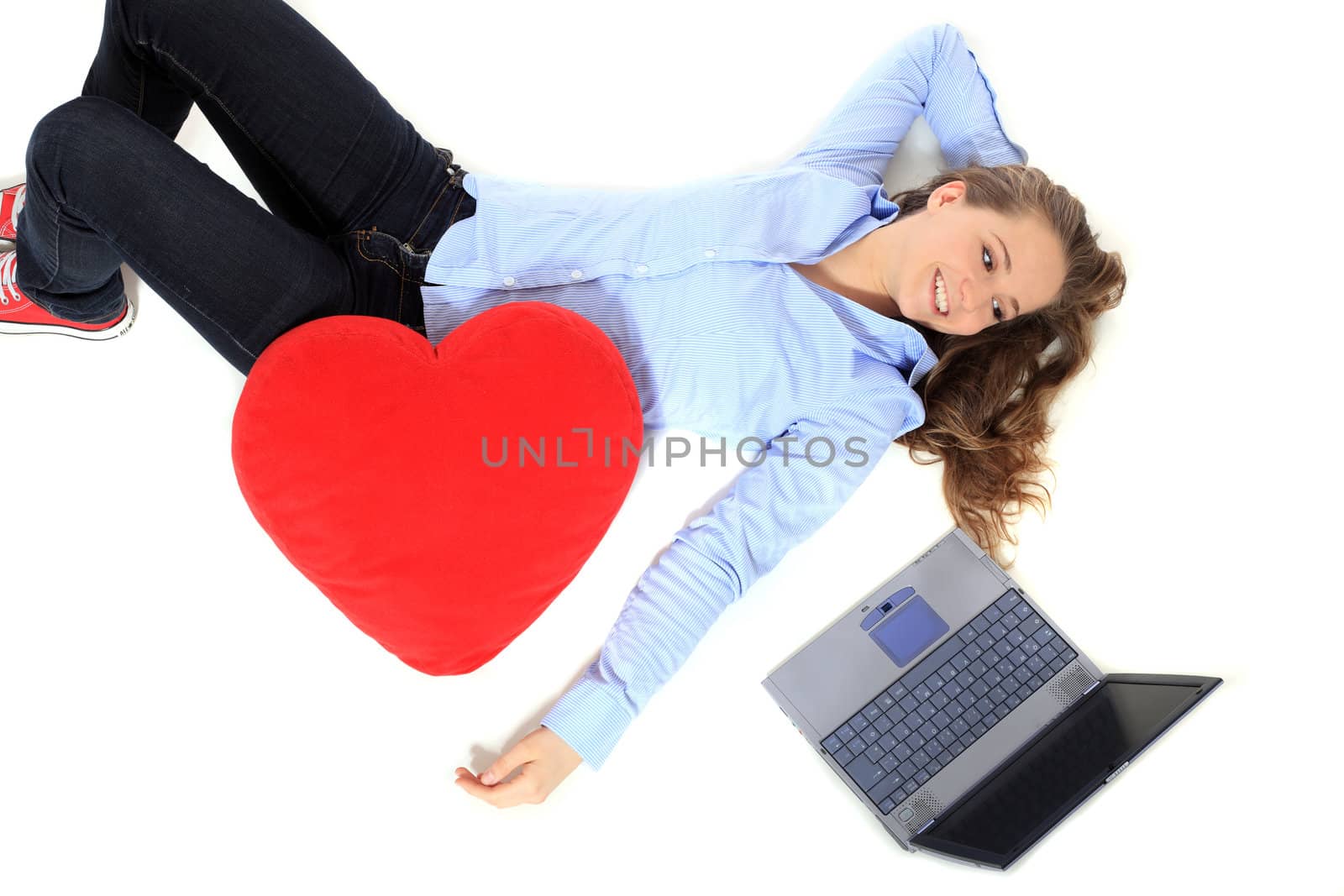 Attractive young girl lying next to her notebook computer holding a heart-shaped pillow. All on white background.