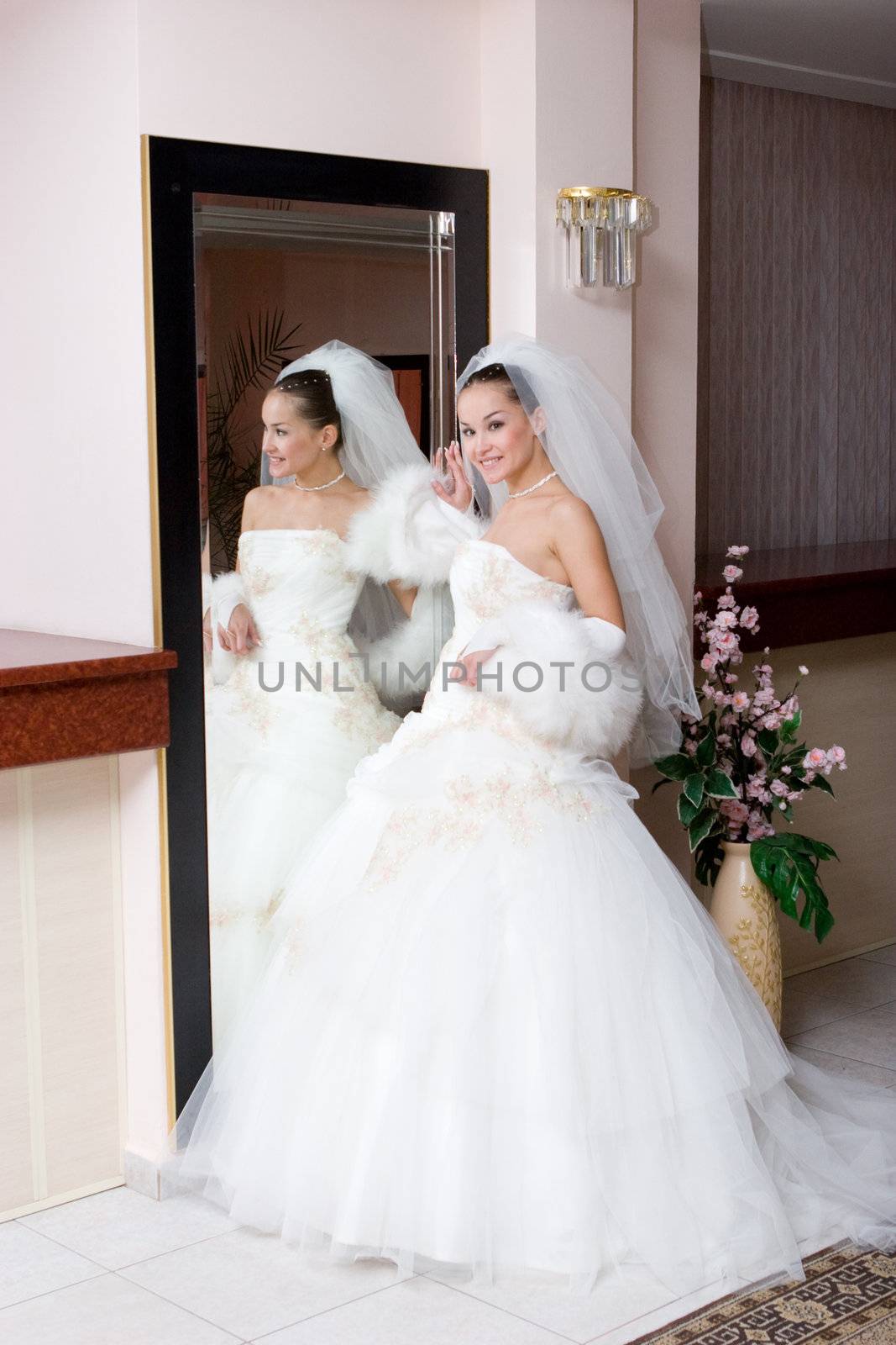a young smiling bride in a beautiful dress stans by a big mirror