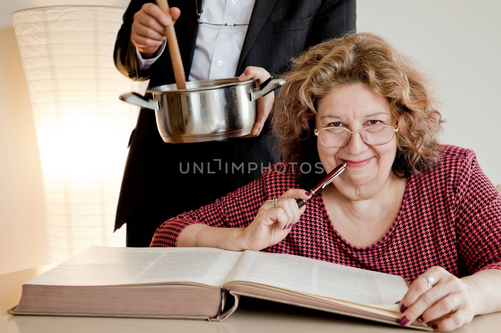 Adult woman smiling at camera over book, man behind her with pot