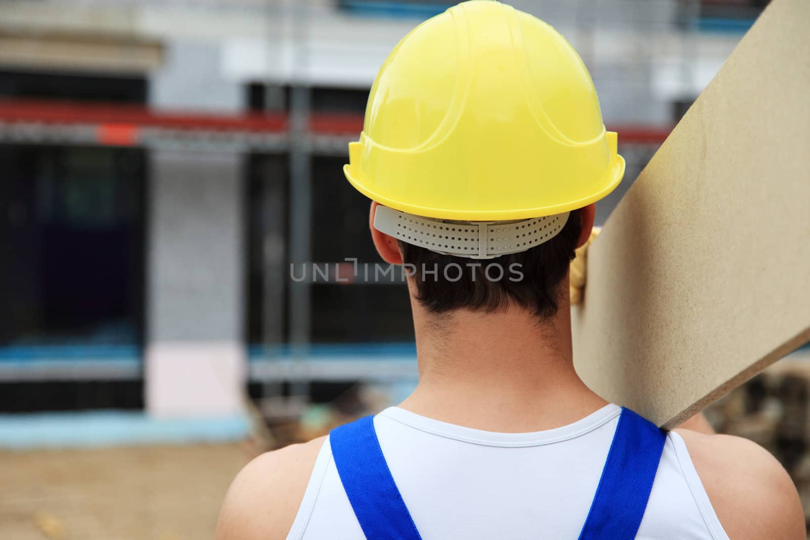 Backview of a manual worker on construction site carrying wooden board.