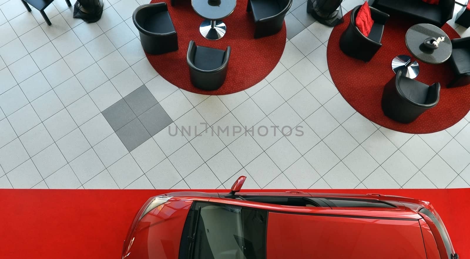 Interior of a car shop with tables and chairs for customers. View from top. Small car standing on red carpet.