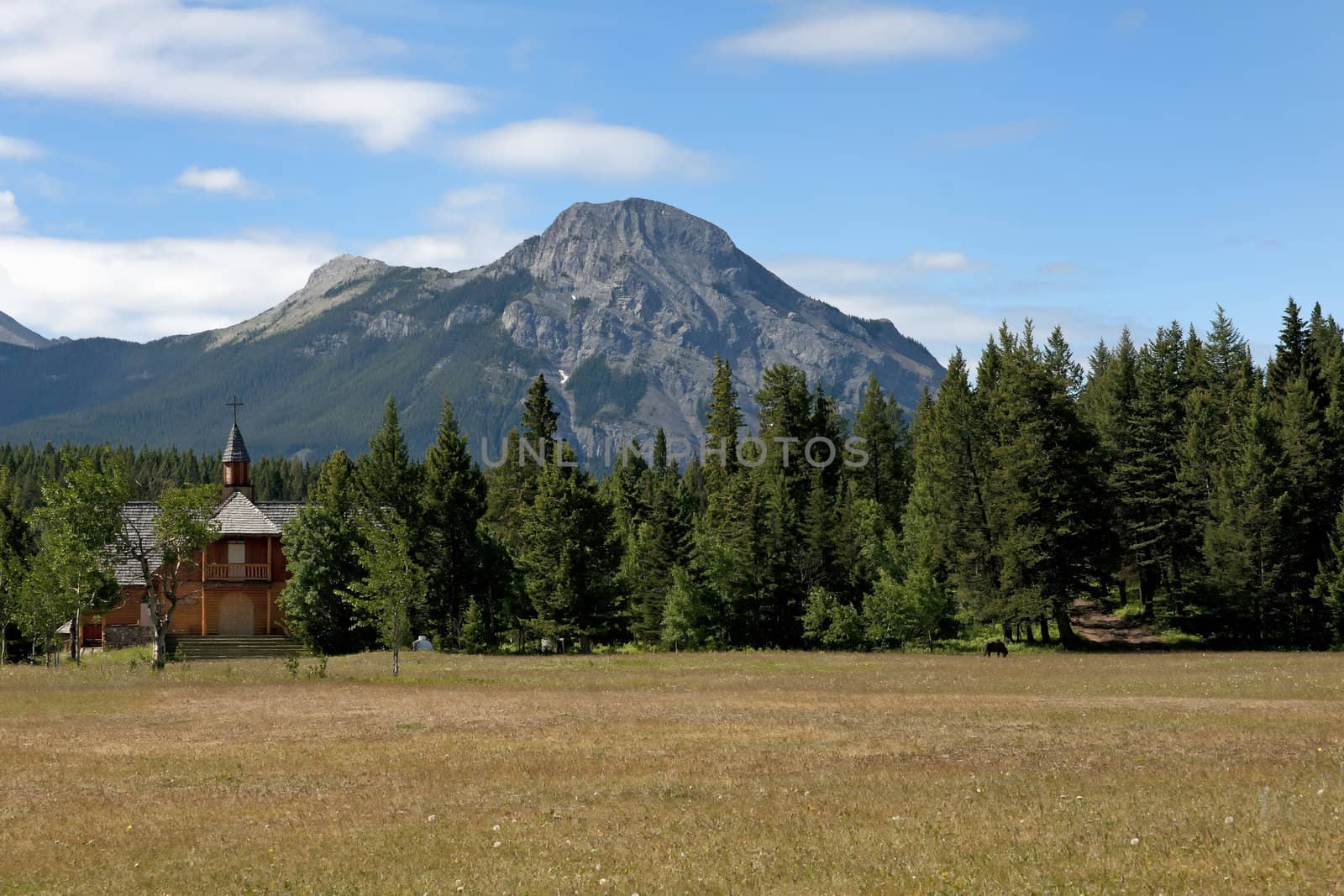 Little red wood church across a plain against forest, rocky moun by Claudine