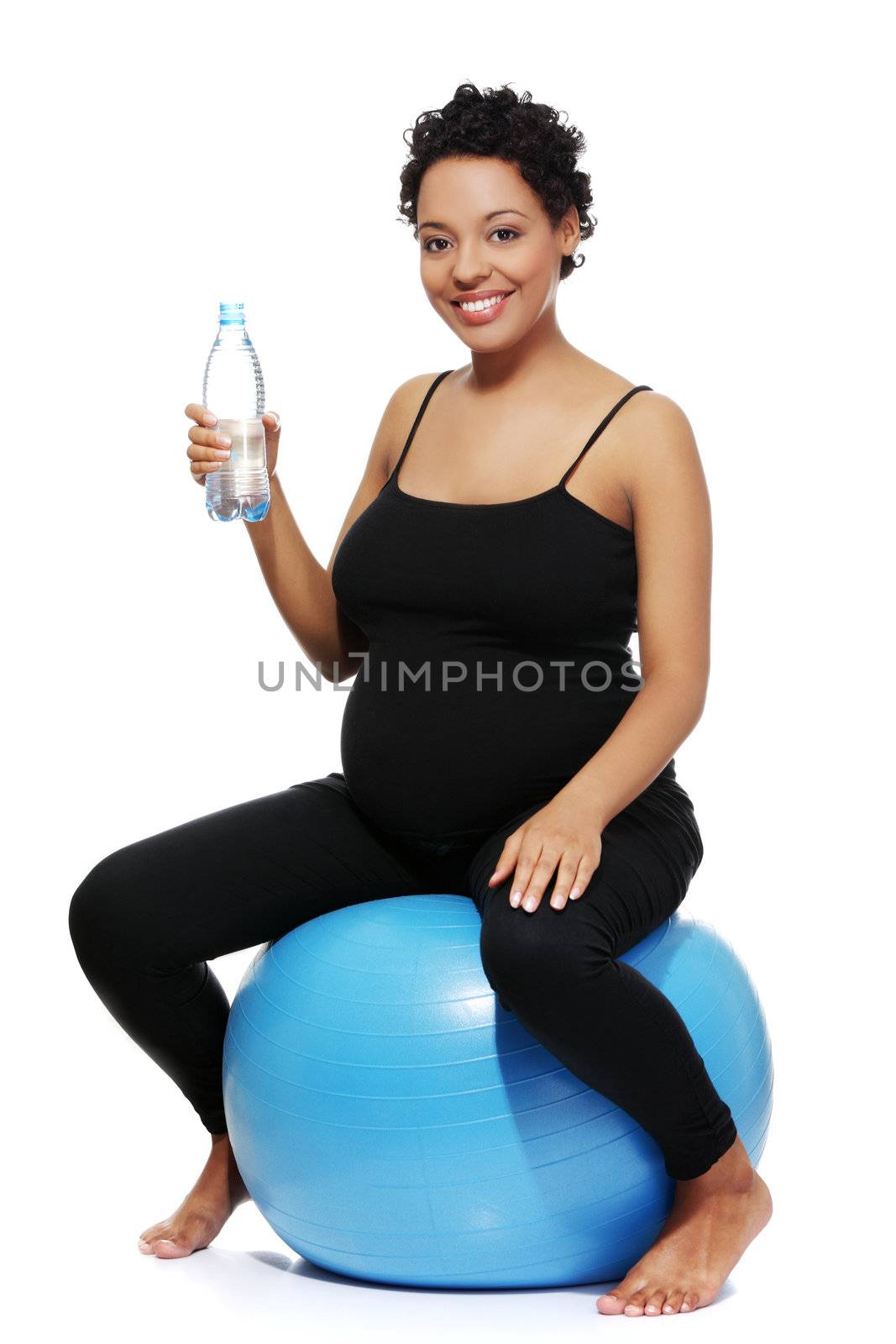 Full lenght portrait of a young smiling pregnant woman sitting on a blue ball, holding a bottle of water, isolated on a white background.