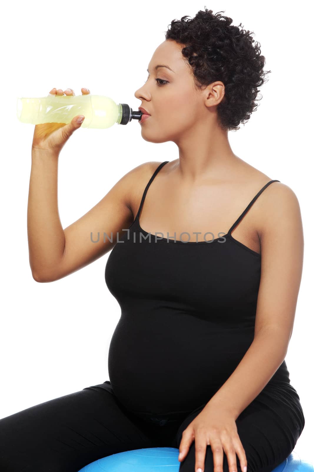 Site view closeup of a young pregnant woman sitting on a blue ball, drinking an energy potion, isolated on a white background.