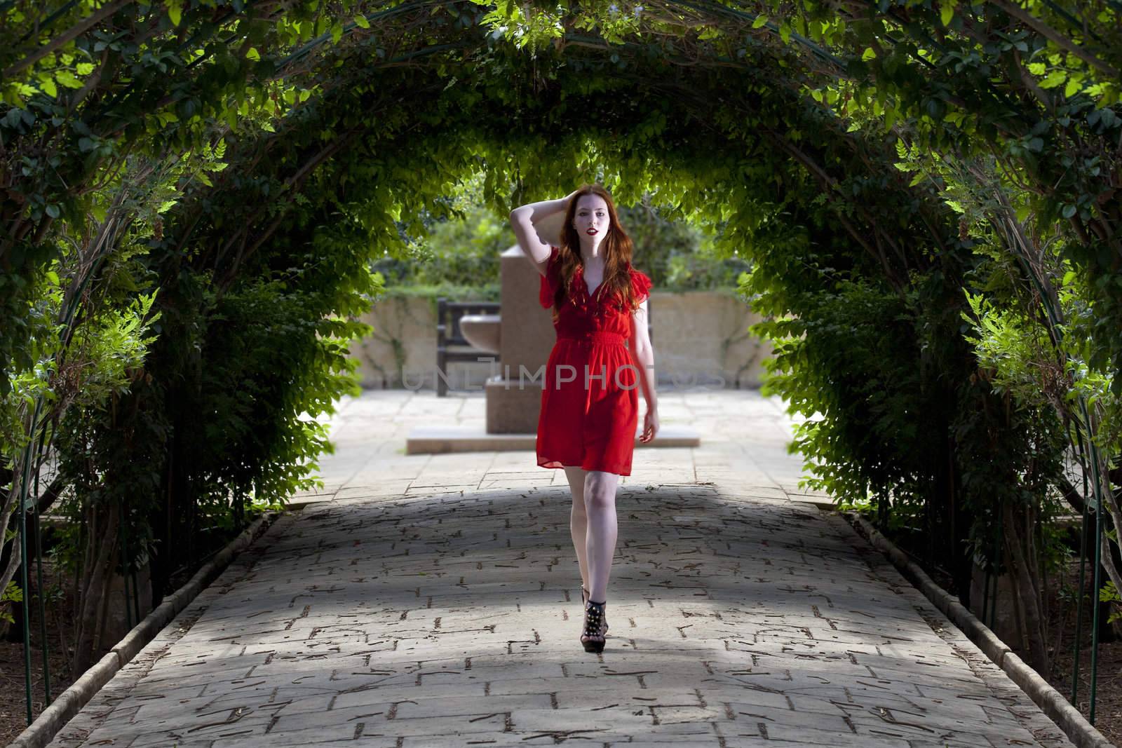 Young woman in red dress walking under arches of foliage