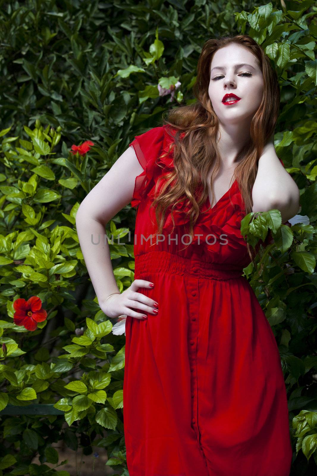 Beautiful model with white skin wearing a red dress with green foliage in background