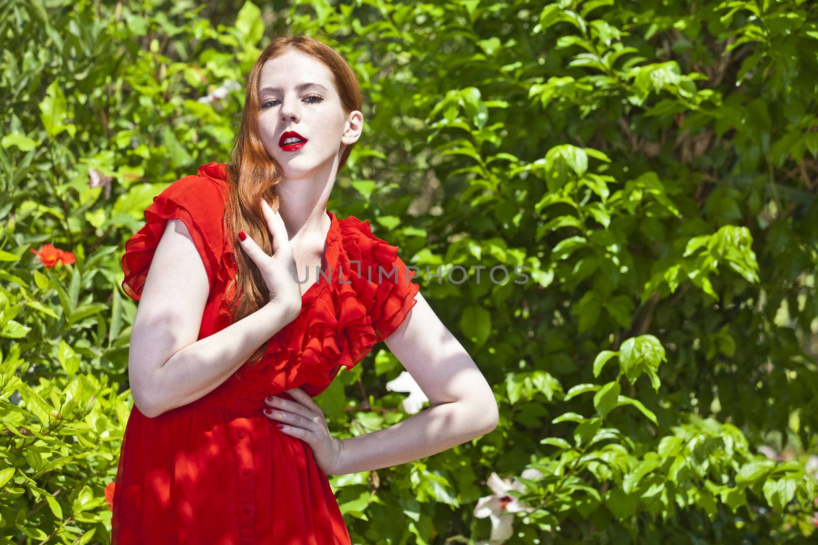 Beautiful model with white skin wearing a red dress with green foliage in background