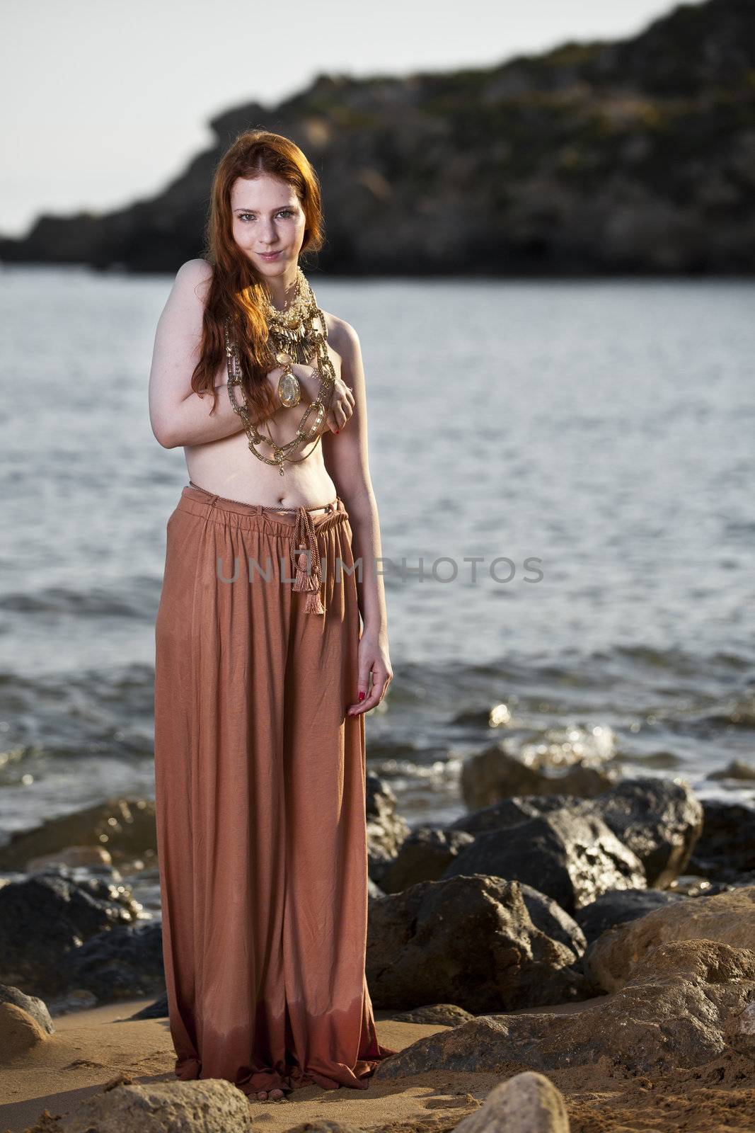 Beautiful woman with pale skin and long red hair wearing Bohemian jewellery and posing semi-nude on a beach