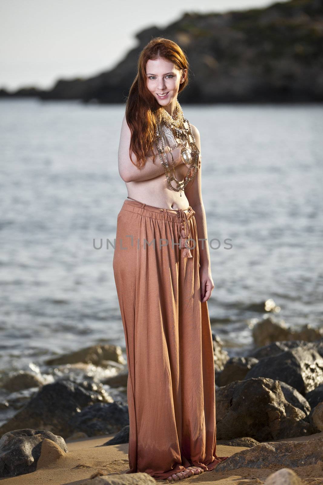 Beautiful woman with pale skin and long red hair wearing Bohemian jewellery and posing semi-nude on a beach