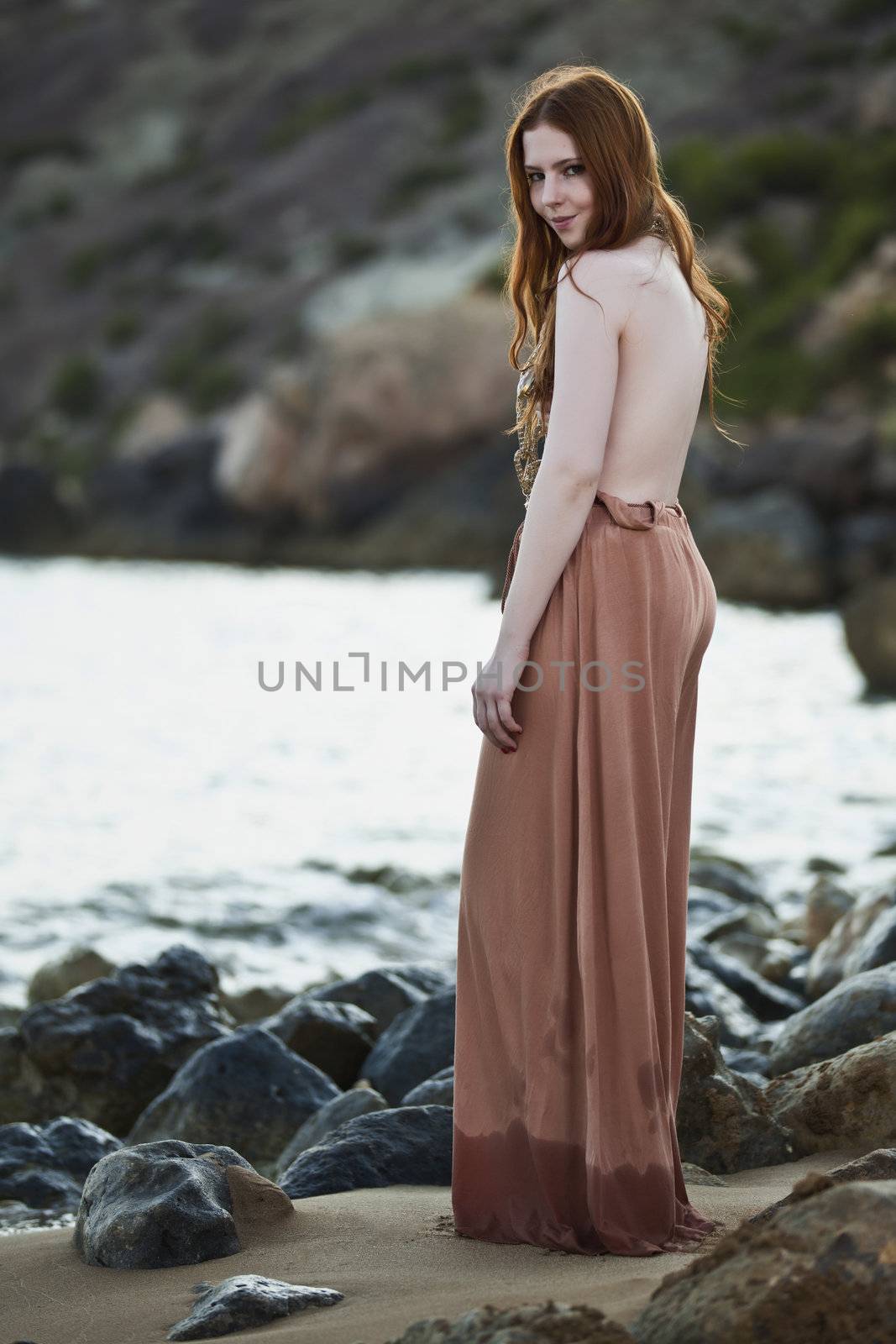 Beautiful woman with pale skin and long red hair wearing Bohemian jewellery and posing semi-nude on a beach