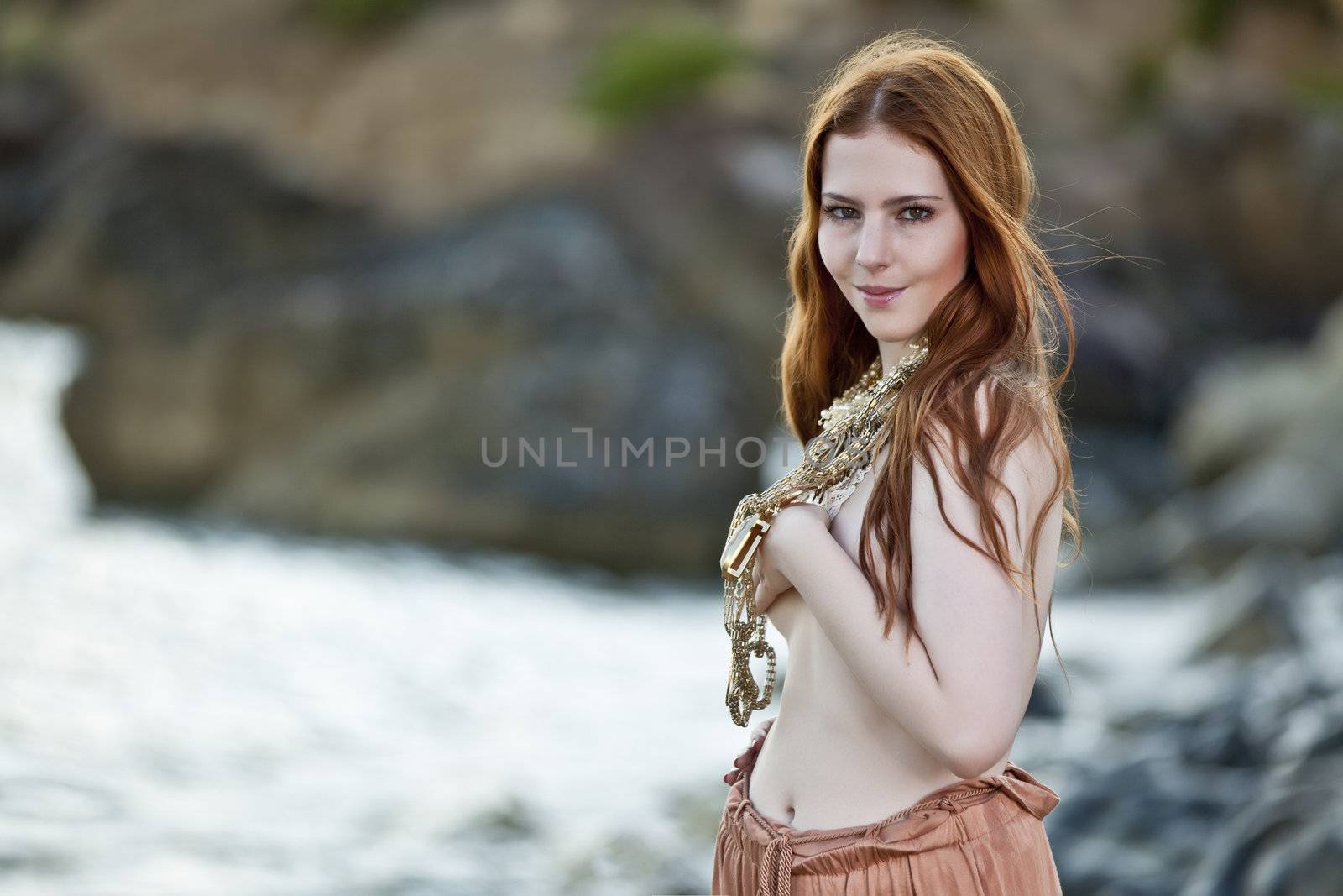 Beautiful woman with pale skin and long red hair wearing Bohemian jewellery and posing semi-nude on a beach