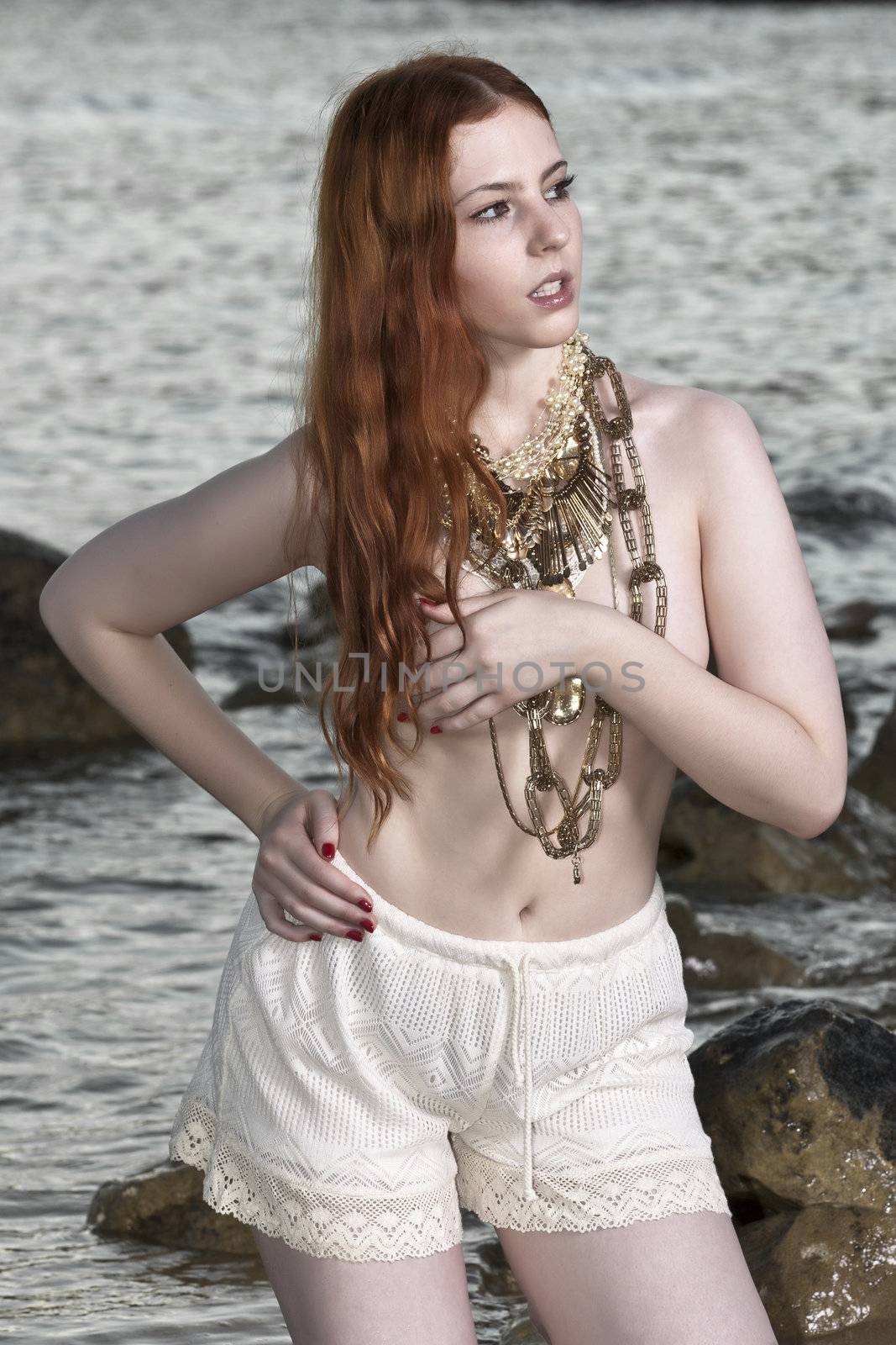 A beautiful woman with pale skin and red hair kneeling on a beach posing semi nude