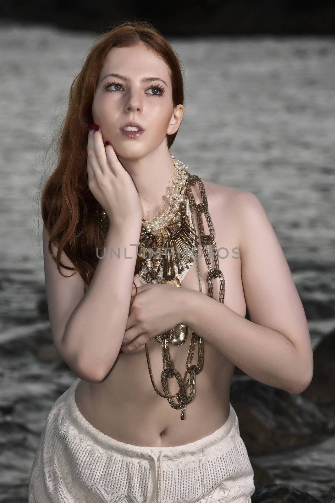 A beautiful woman with pale skin and red hair kneeling on a beach posing semi nude