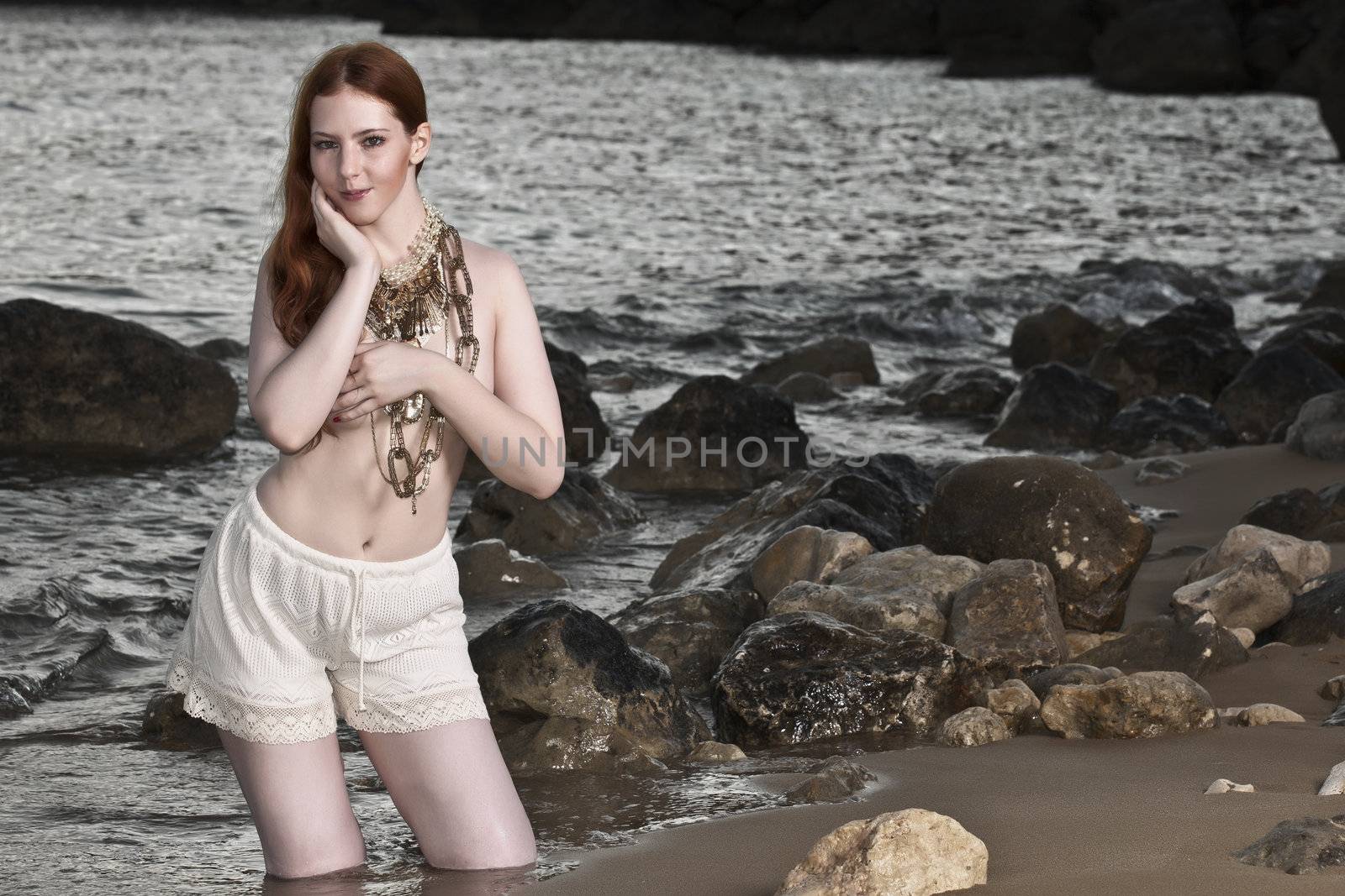 A beautiful woman with pale skin and red hair kneeling on a beach posing semi nude