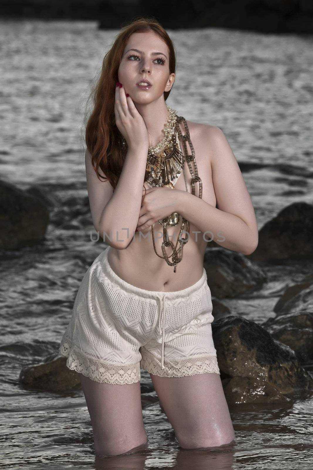 A beautiful woman with pale skin and red hair kneeling on a beach posing semi nude