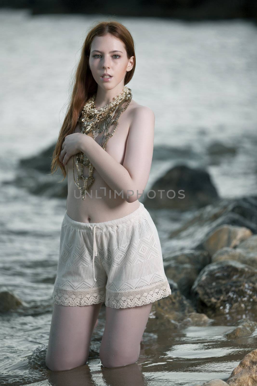 A beautiful woman with pale skin and red hair kneeling on a beach posing semi nude