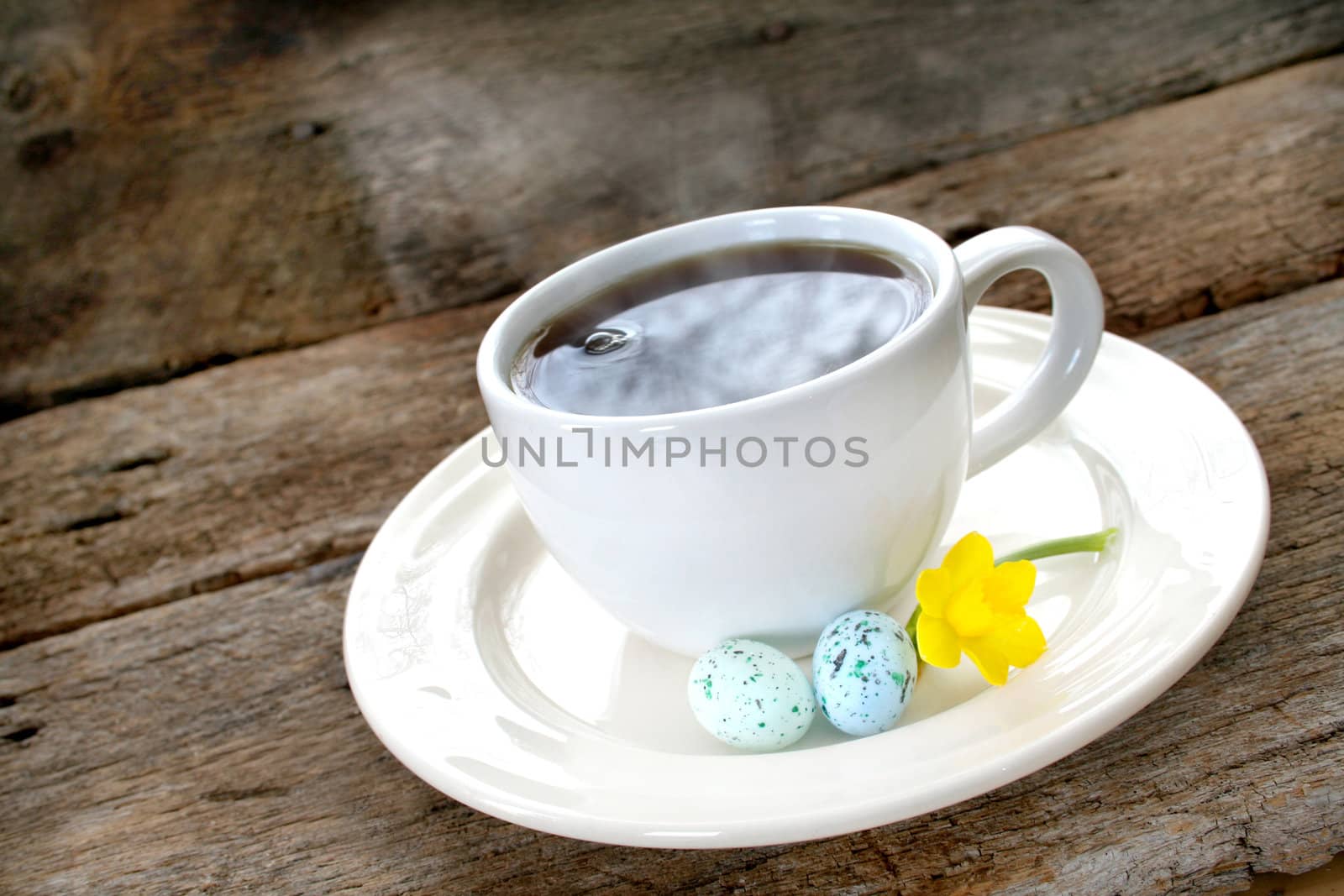 Cup of coffee sitting on a saucer with Easter eggs and a daffodil.  Steam can be seen coming out of the cup of hot coffee.