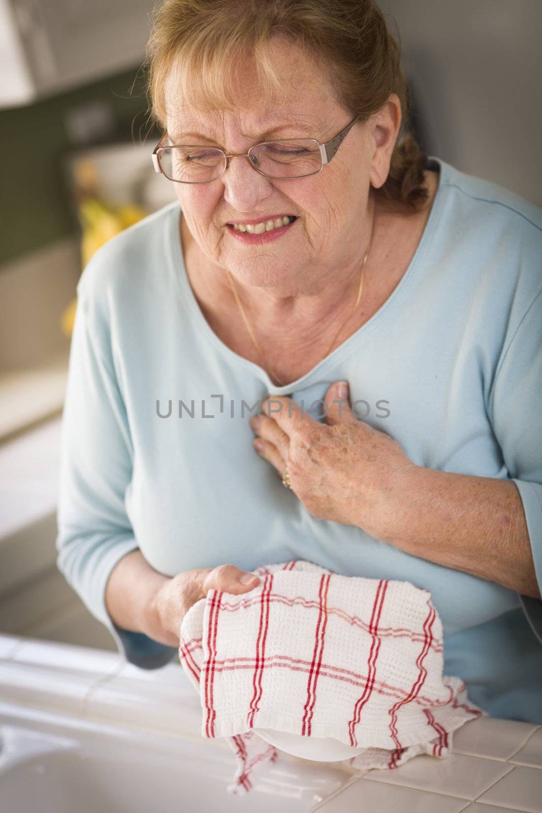 Grimacing Senior Adult Woman At Kitchen Sink With Chest Pains.