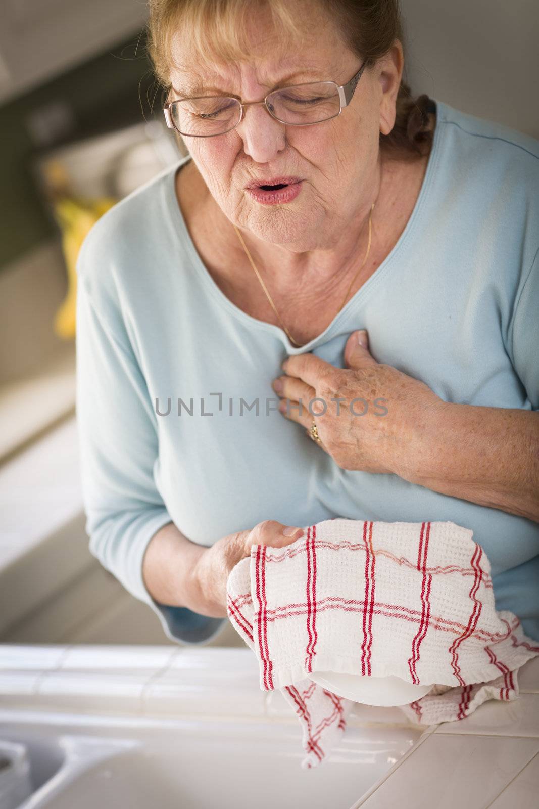 Grimacing Senior Adult Woman At Kitchen Sink With Chest Pains.