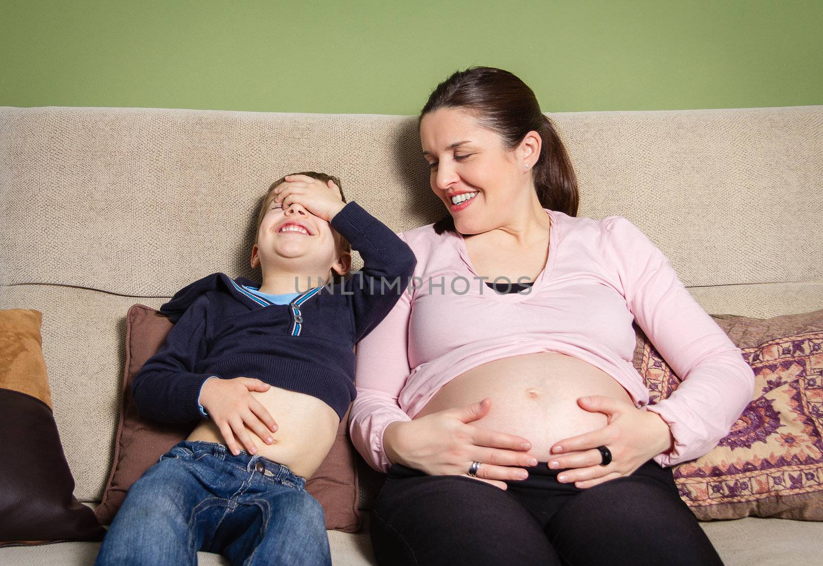 Pregnant mother and son laughing when comparing their bellies sitting on a sofa