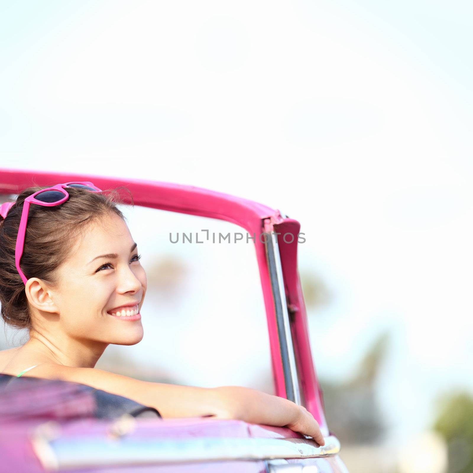 Car woman smiling happy enjoying car road trip travel vacation. Young retro woman in pink vintage convertible car looking to side at copy space. Beautiful young multiracial caucasian / chinese asian female model. Photo from Havana, Cuba.