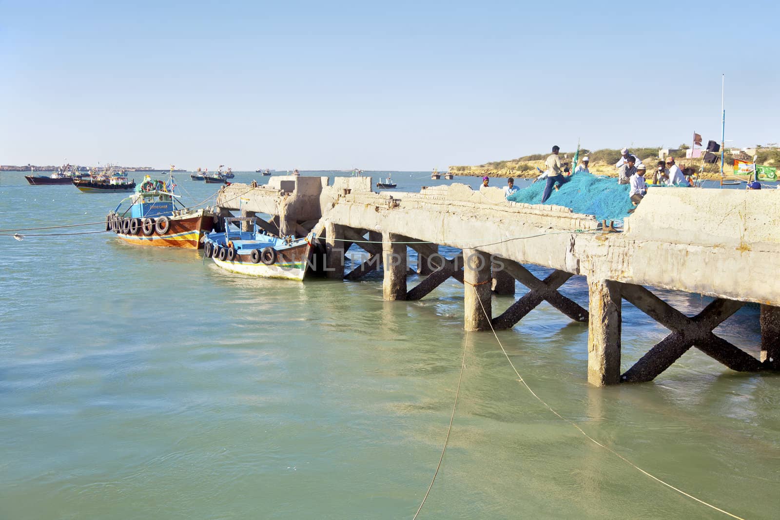 Dwarka Roadtrip. Landcsape capture of fishnet menders taking a tea break at Bet Dwarka pier. Moored and anchored boats in the barhor under a clear blue hazy sky completes an idyllic picturesque scene that separates the mainland from the Island