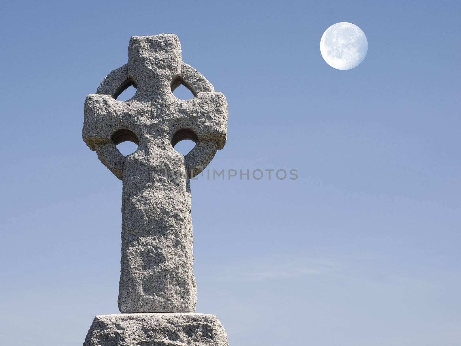 Celtic cross in a cementary with a full moon in the blue sky.