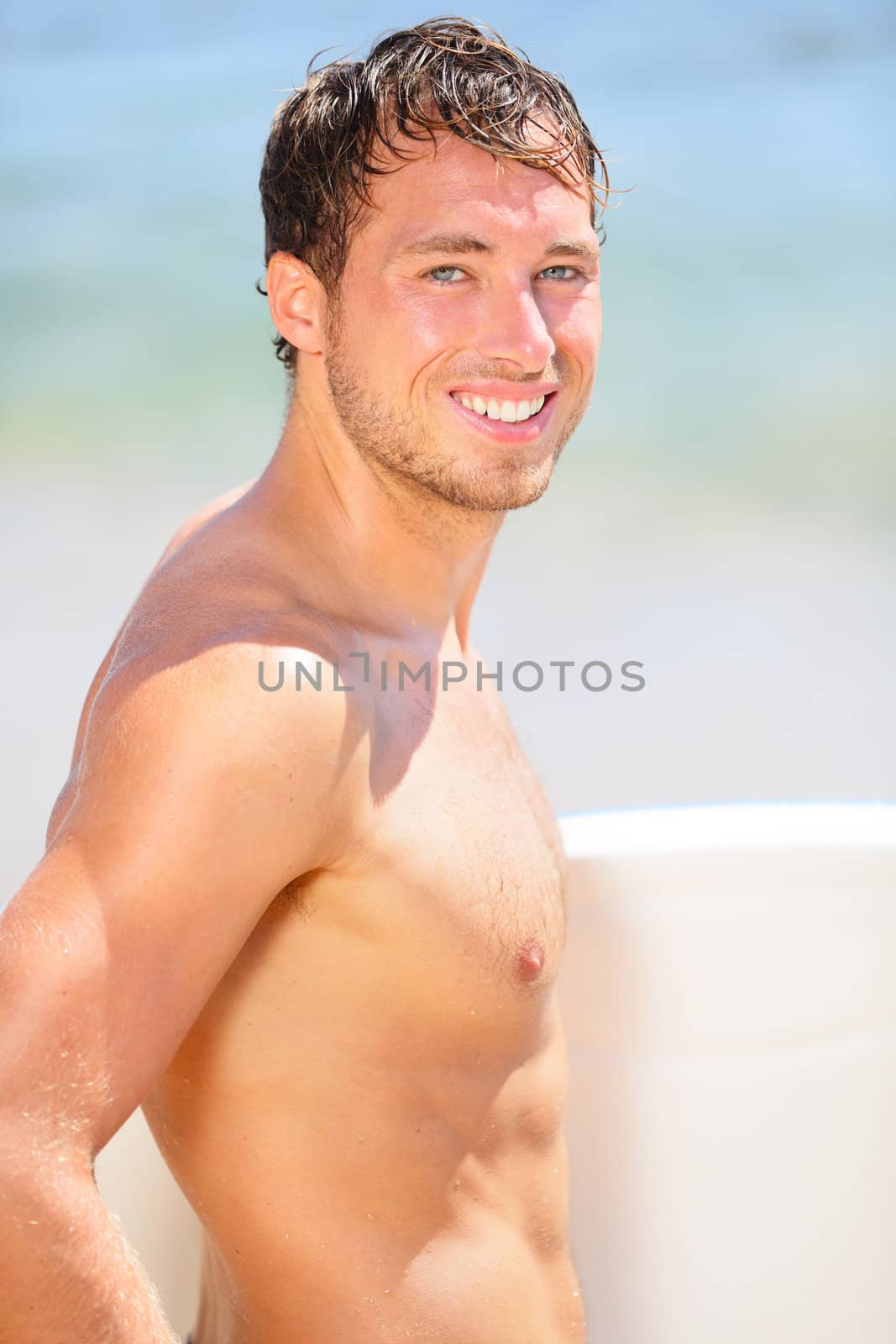 Surfer beach man portrait. Happy handsome young male beach surfing model smiling at camera holding surfboard under the sun on sunny summer day during holidays vacation. Good looking guy in his 20s.