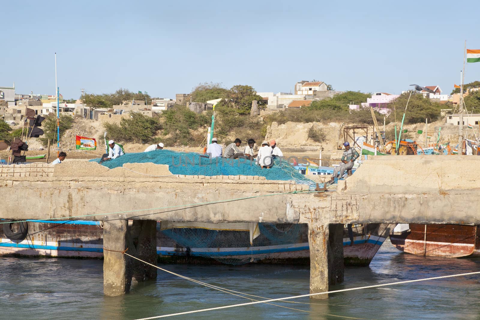 Landscape capture of fishnet menders hard at work under the hot sun at Bet Dwarka pier. Moored and anchored boats in the harbor under a clear blue hazy sky completes an idyllic picturesque scene that separates the mainland from the Island