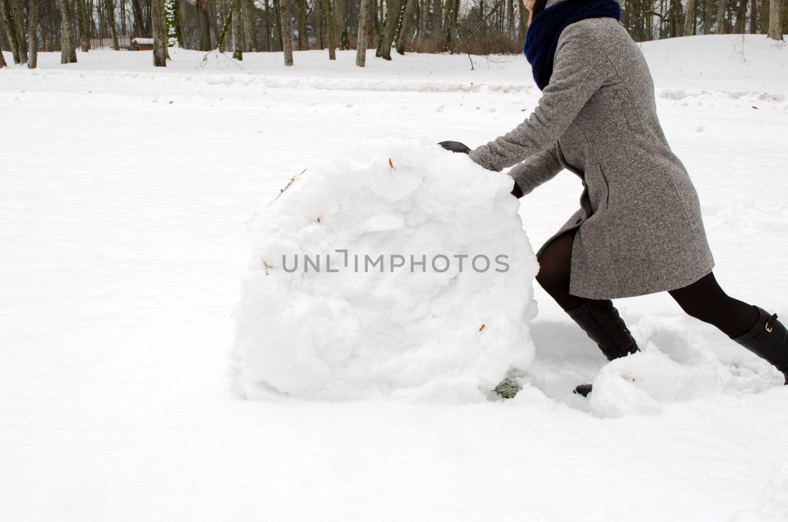 woman in grey coat push big snow ball roll in winter meadow. snowman building.