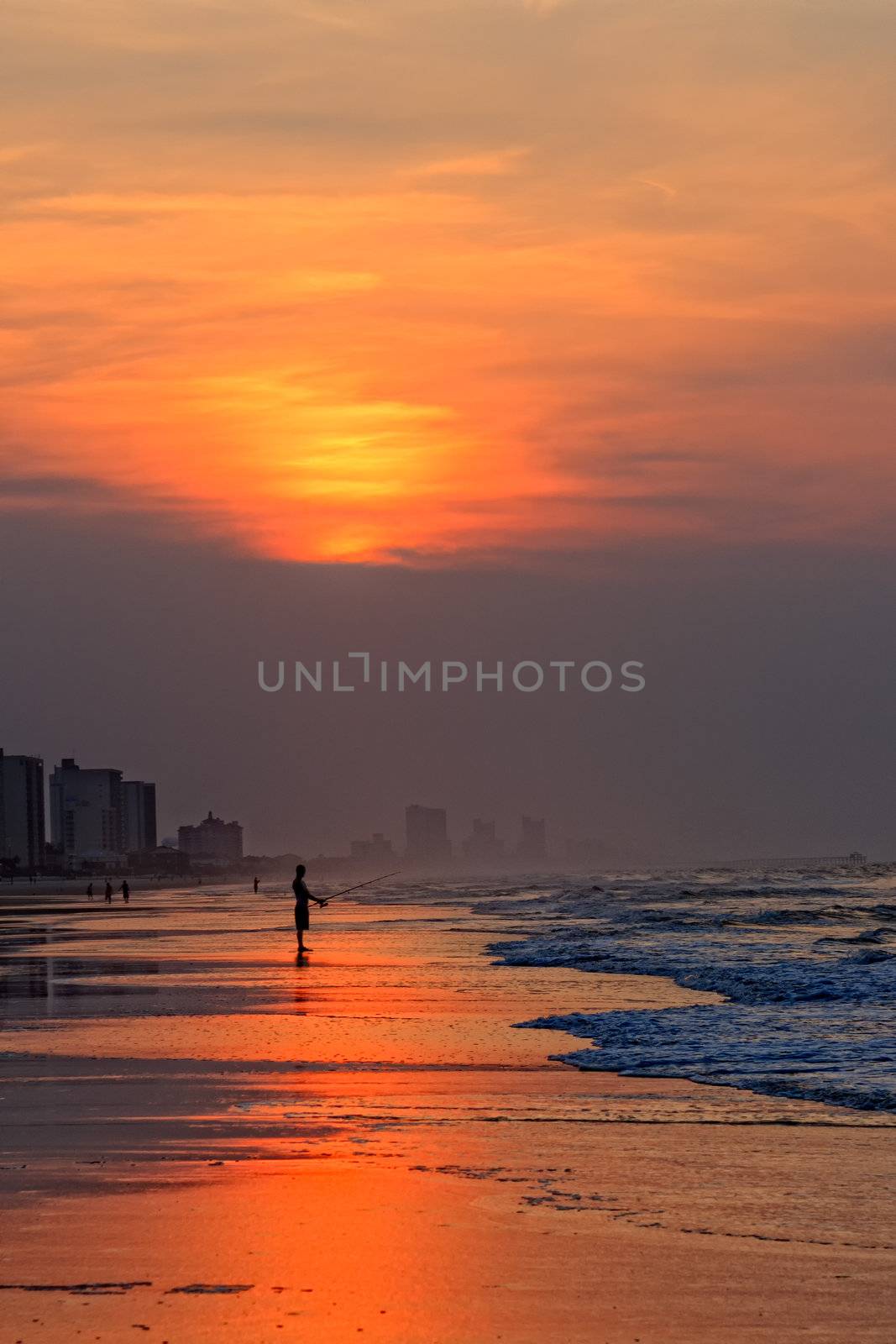 A silhouette of a lone fisherman fishing in the early morning in the Atlantic Ocean from the beach as the sun rises.