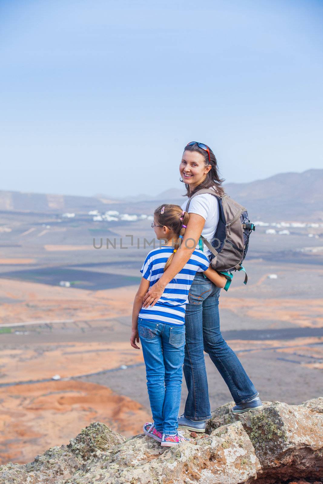 Young mother and daughter with backpack standing on cliff's edge and looking to a sky. Vertical view