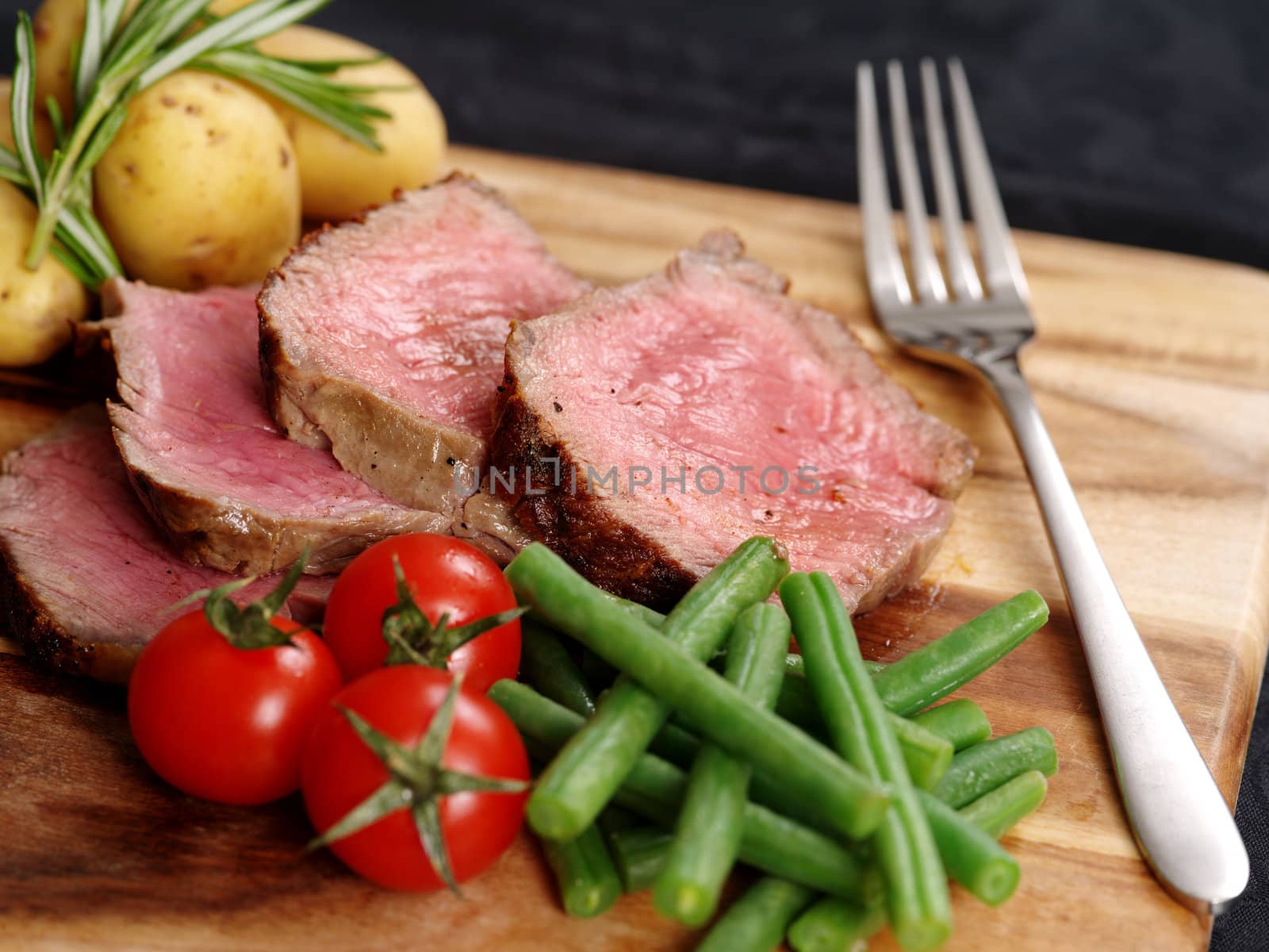 Photo of steak dinner with thick slices of sirloin, cherry tomatoes, green beans and potatoes on a wooden board.