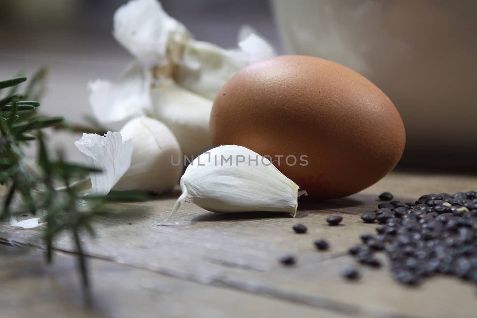 Close up detail of some kitchen ingredients comprising of a brown egg, garlic, fresh rosemary and black lentils. All set on a landscape format against a wooden background.