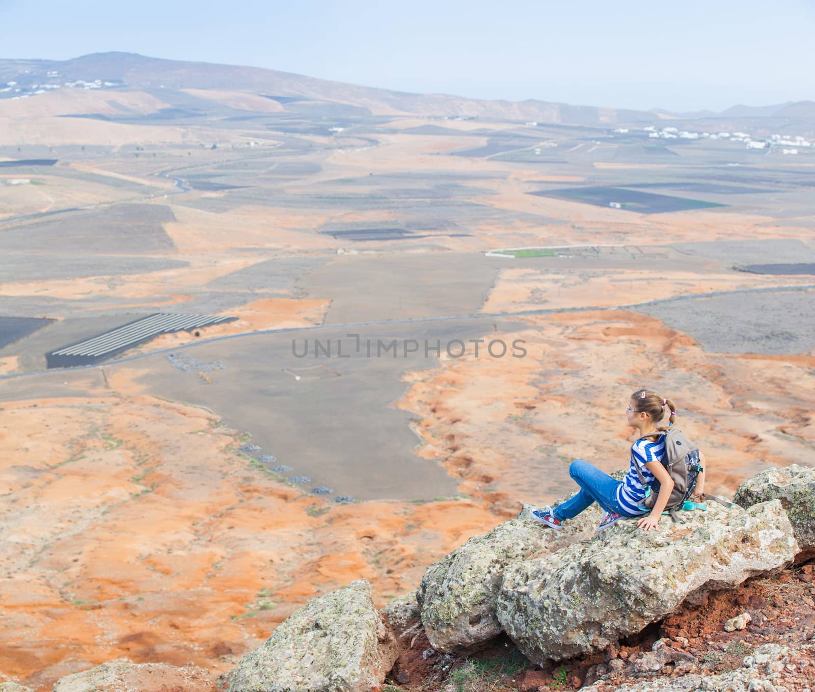 Young girl sitting with backpack on cliff's edge and looking to a sky. Lanzarote
