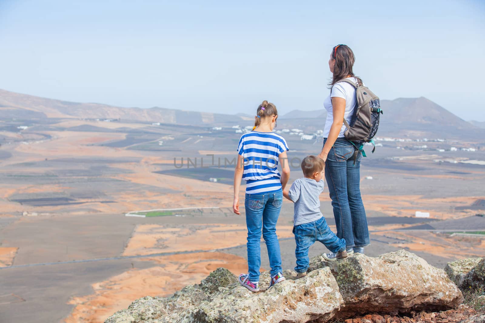 Young mother with backpack and her chid standing on cliff's edge and looking to a sky