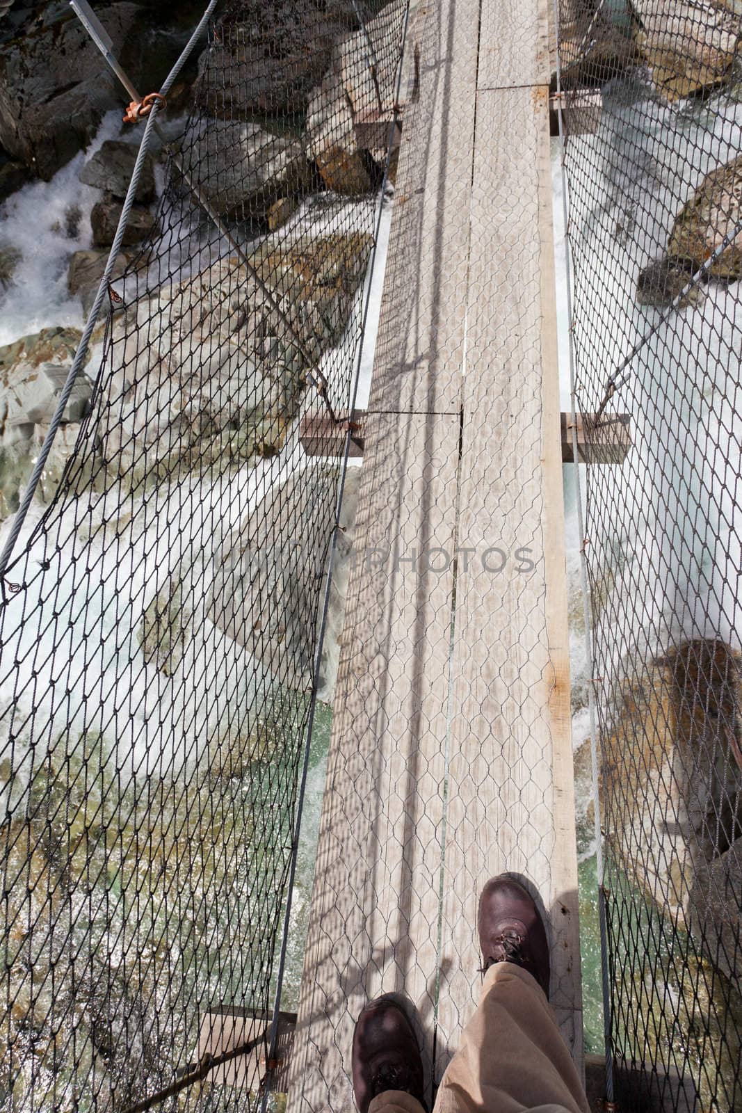 Booted feet of man walking on narrow swing bridge over clear fresh white water rushing through a rocky gorge