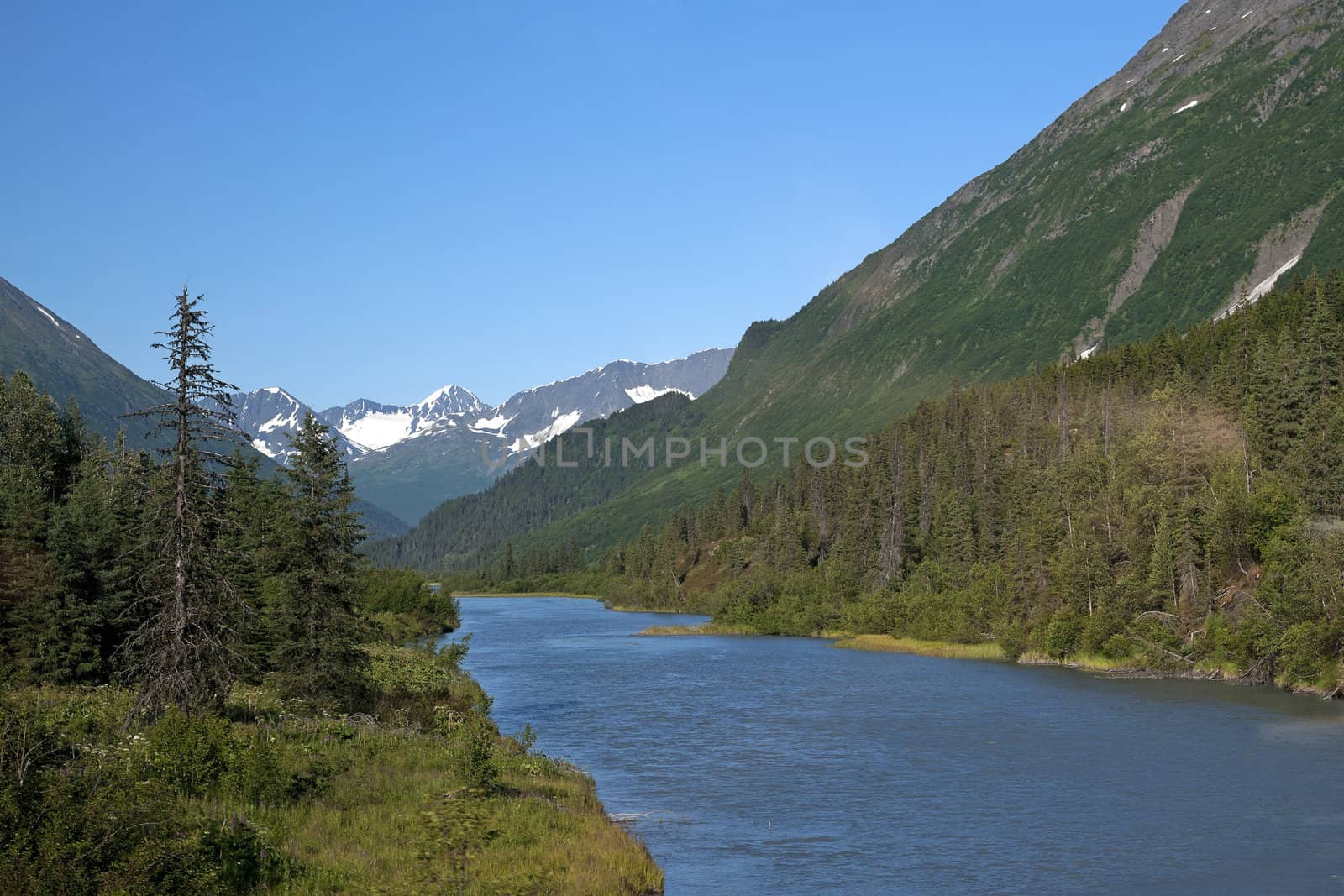 Blue water valley cuts through green mountains with snow patches by Claudine