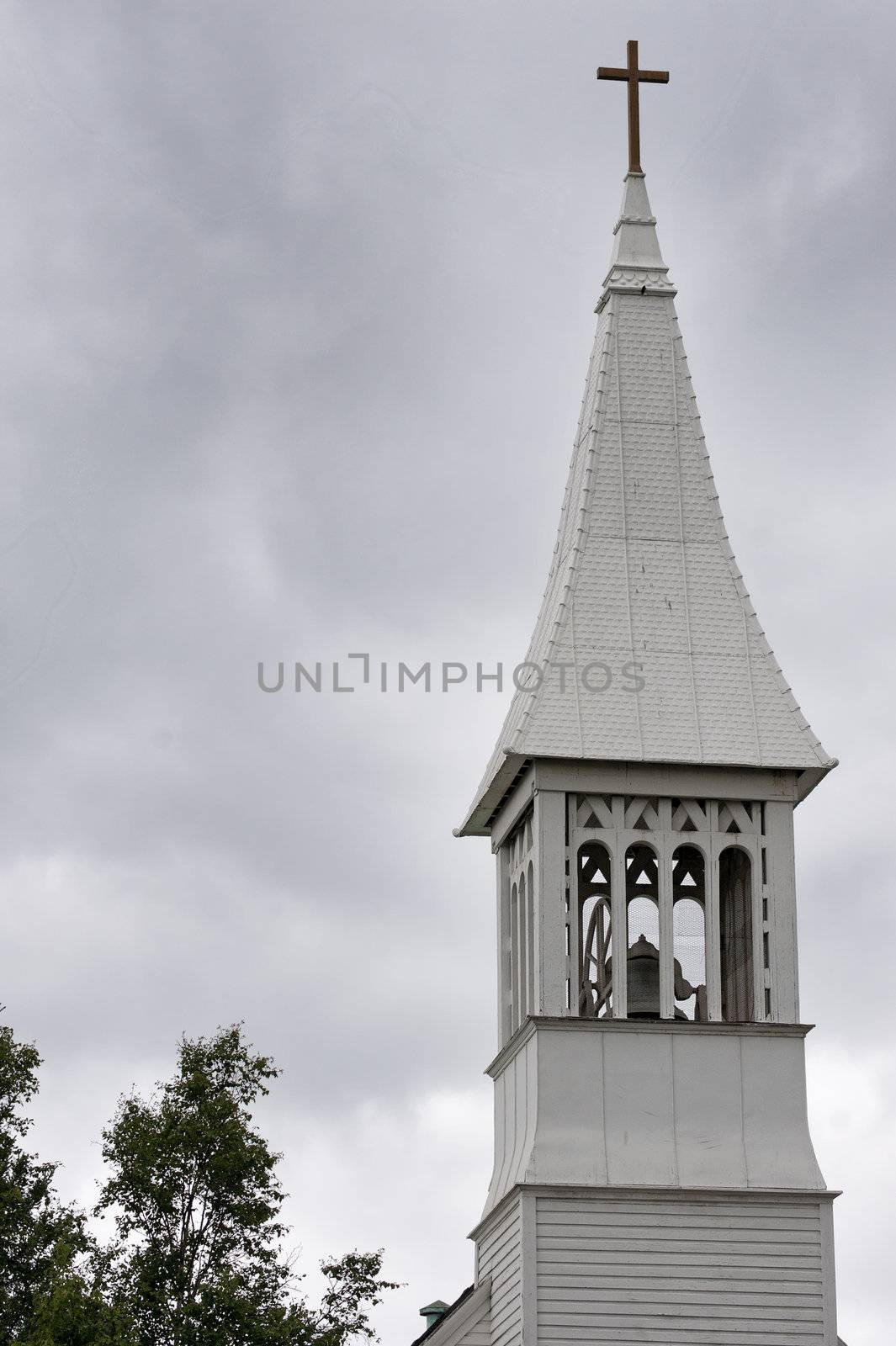 White tower with cross and bell against grey skies.