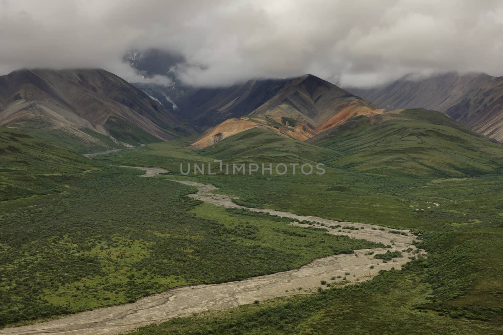 Muddy river meanders down from colorful mountains under stormy s by Claudine