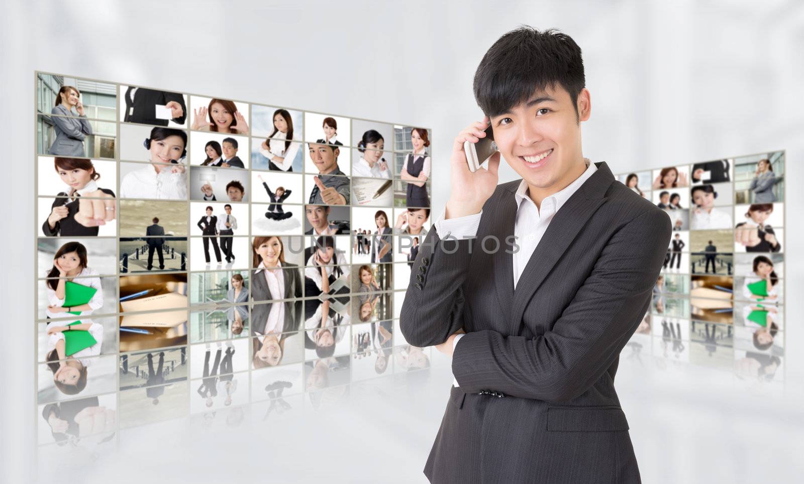 Young businessman talk on phone and stand in front of tv screen wall showing pictures of business concept by Asian business people. Concepts about business, communication, teamwork or social network.