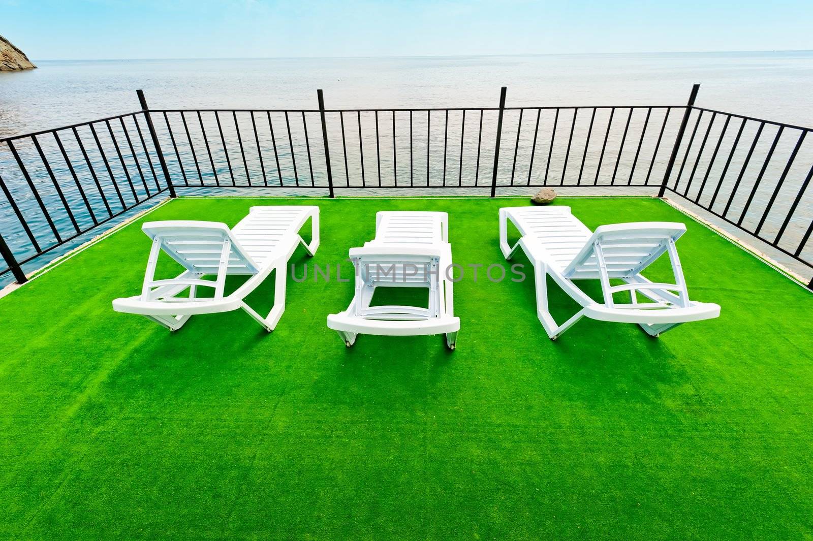 Three white beach lounger on a green deck with a view of the sea.