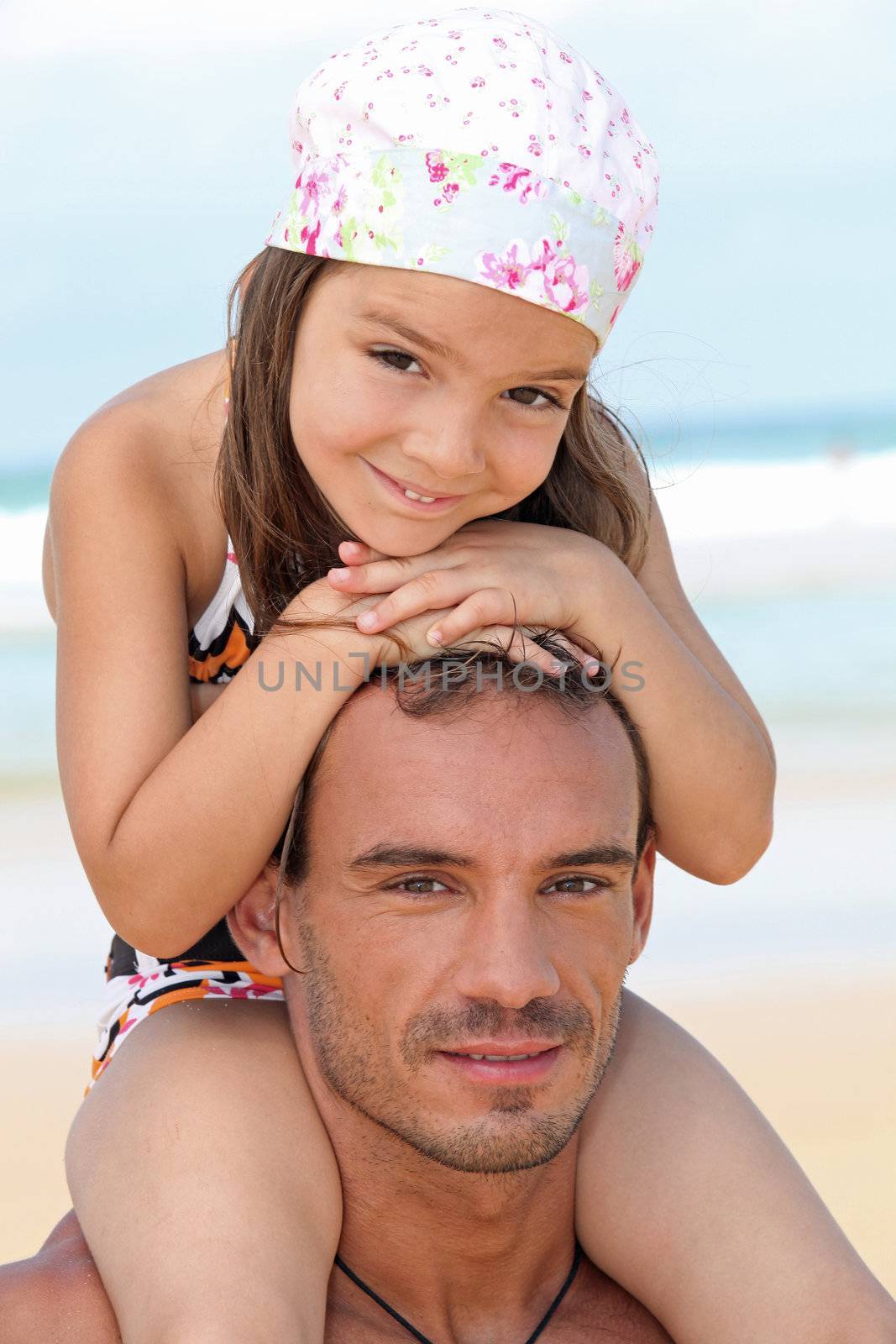 little girl sitting on father's shoulders on the beach by phovoir