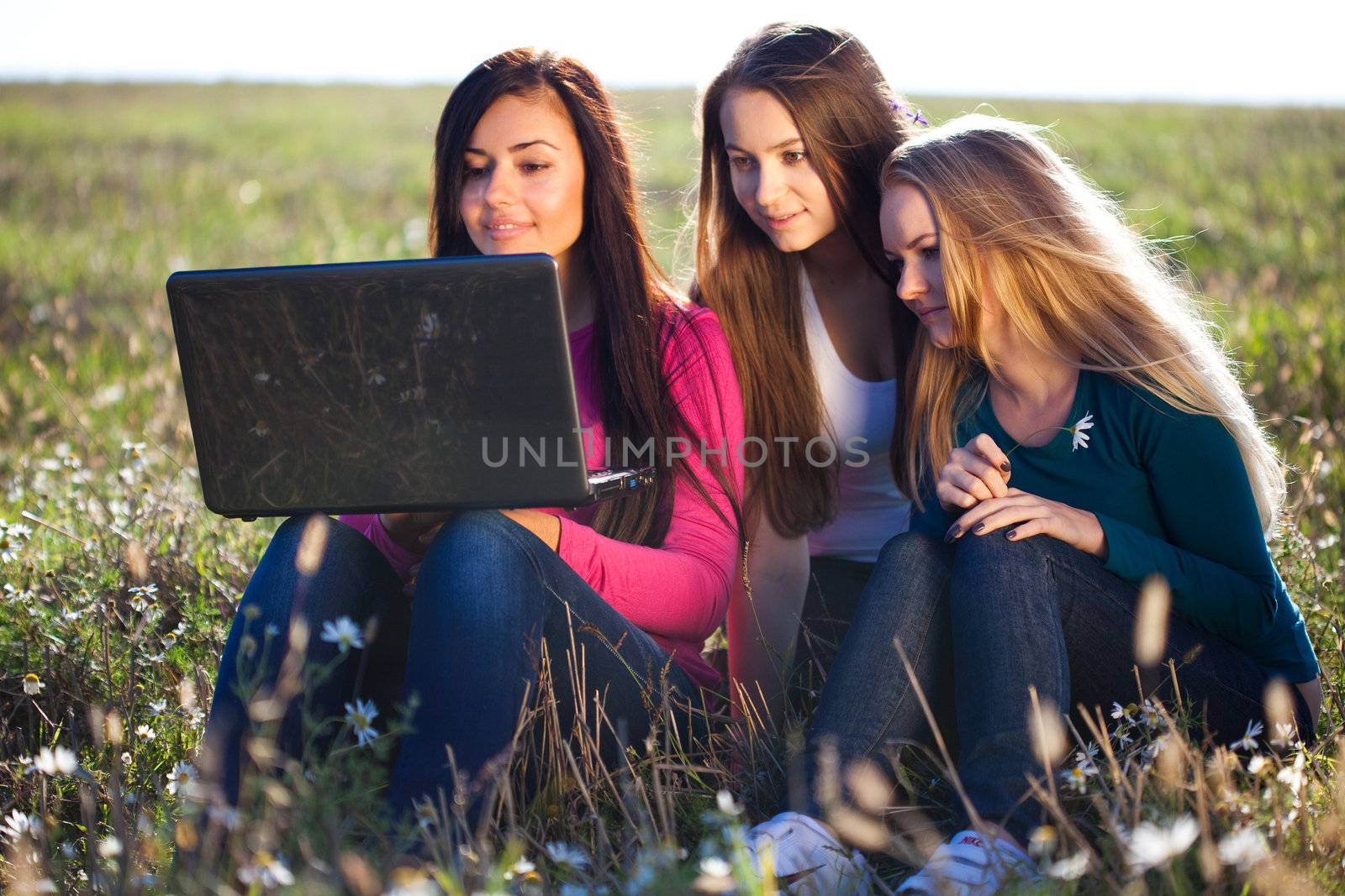 three young beautiful woman with a laptop sitting in the field o by jannyjus